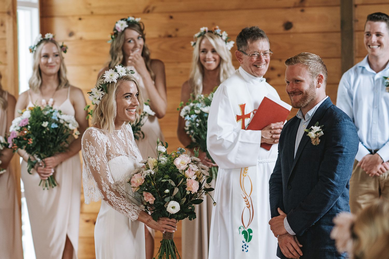 Wedding party in white and neutral tones poses together in rustic wooden venue with floral decorations and natural light.