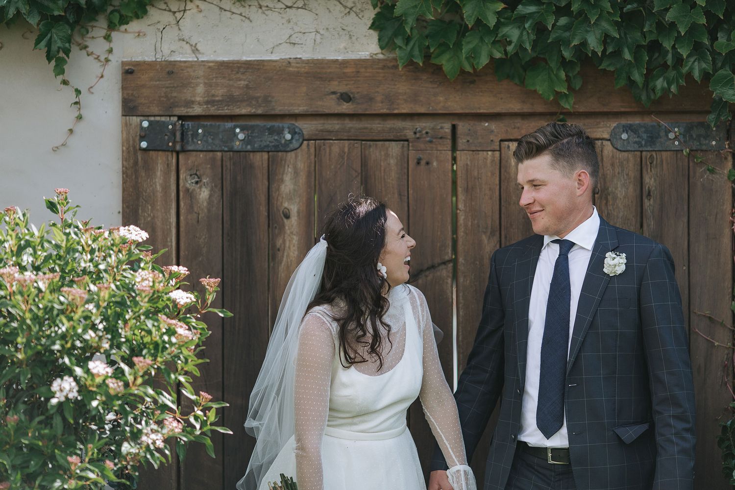 A couple stands together in a lush garden setting against a rustic wooden fence with climbing vines and flowers.