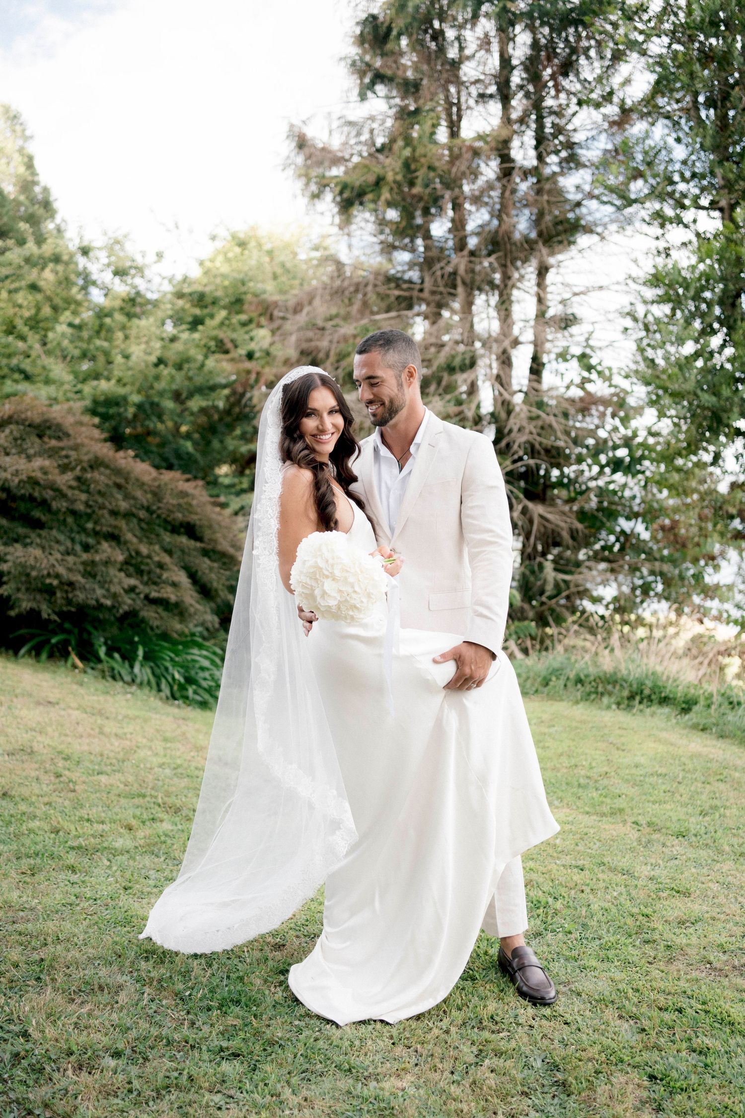 Newlyweds in all-white attire pose together on a grassy hillside surrounded by trees.