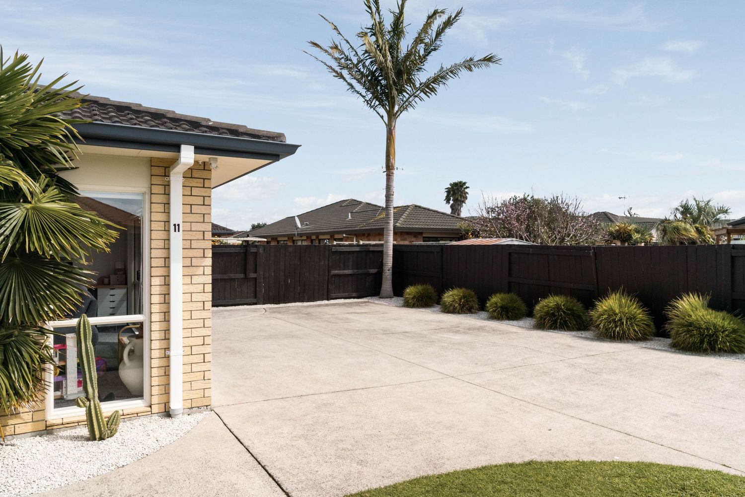Modern house exterior with concrete driveway flanked by landscaped gardens and a young palm tree in suburban setting.