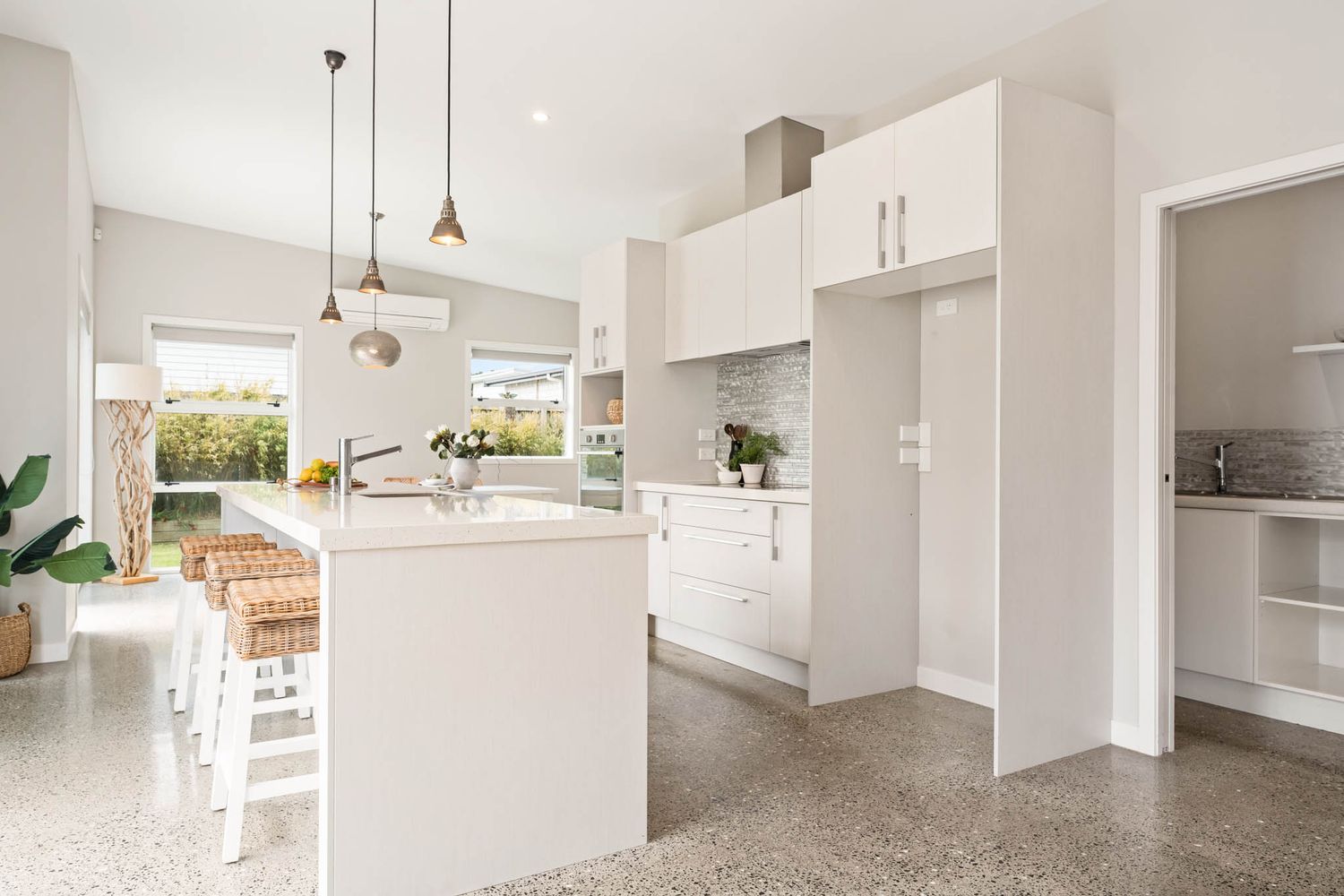 Modern white kitchen with polished concrete floors, island bench, and pendant lighting opens to outdoor area.