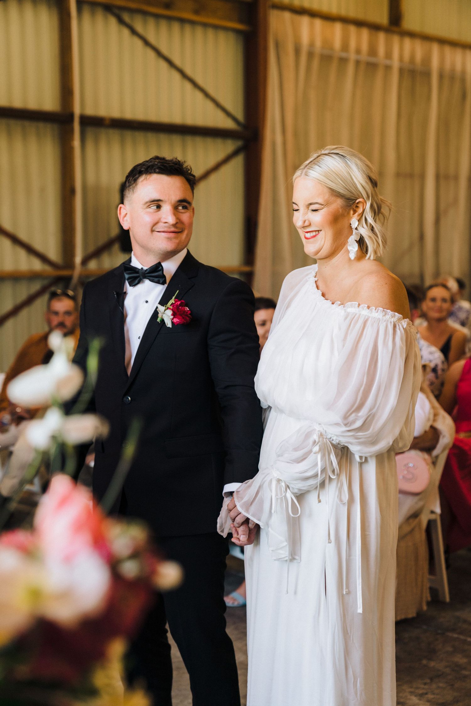 Wedding ceremony in rustic venue with couple in white dress and black tuxedo surrounded by guests.