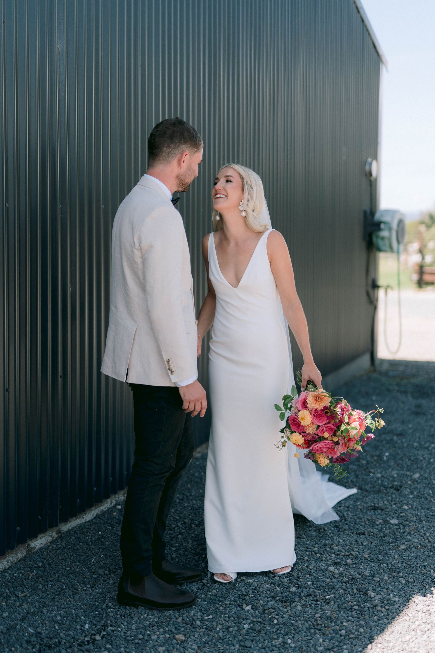 A couple in wedding attire share an intimate moment against a dark wall outdoors.