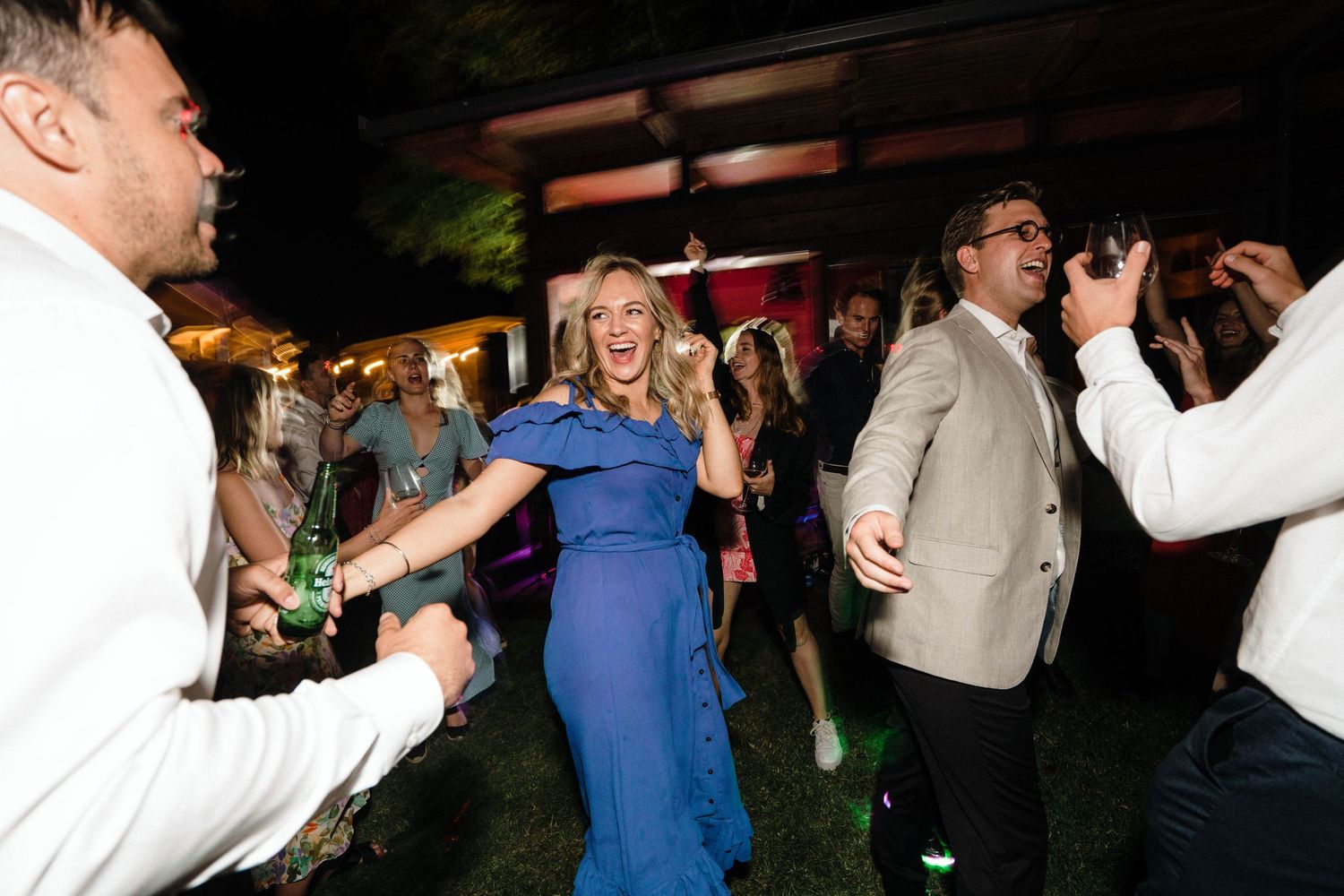 Partygoers in formal attire dance enthusiastically at a nighttime event with a person in a blue dress at center.