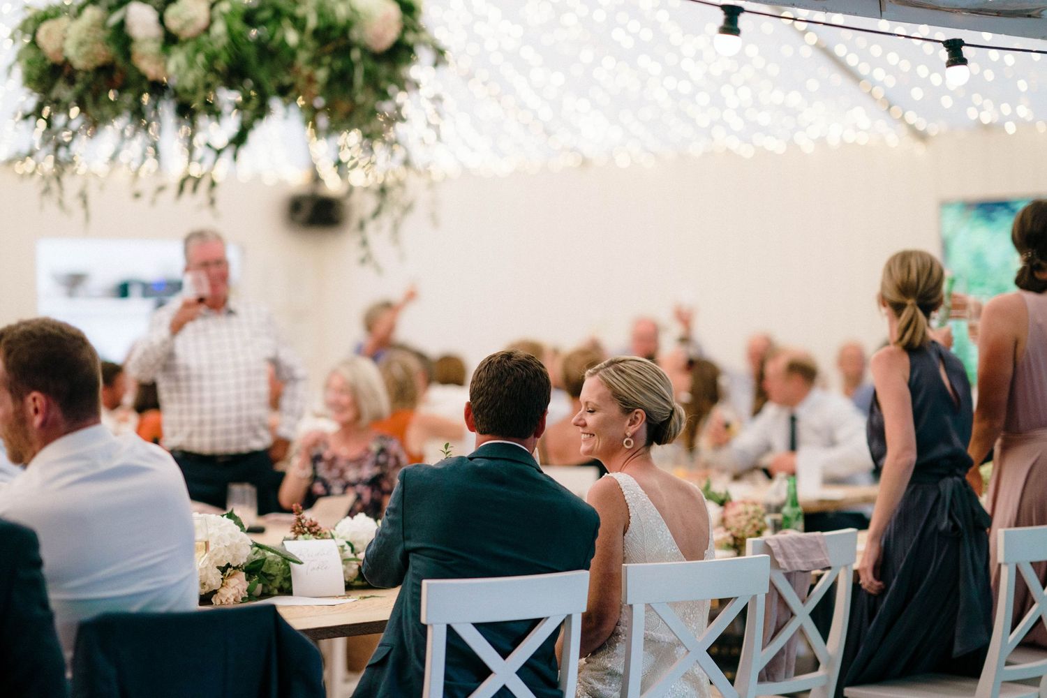 Wedding reception setup with white cross-back chairs and hanging lights creating an elegant outdoor atmosphere.