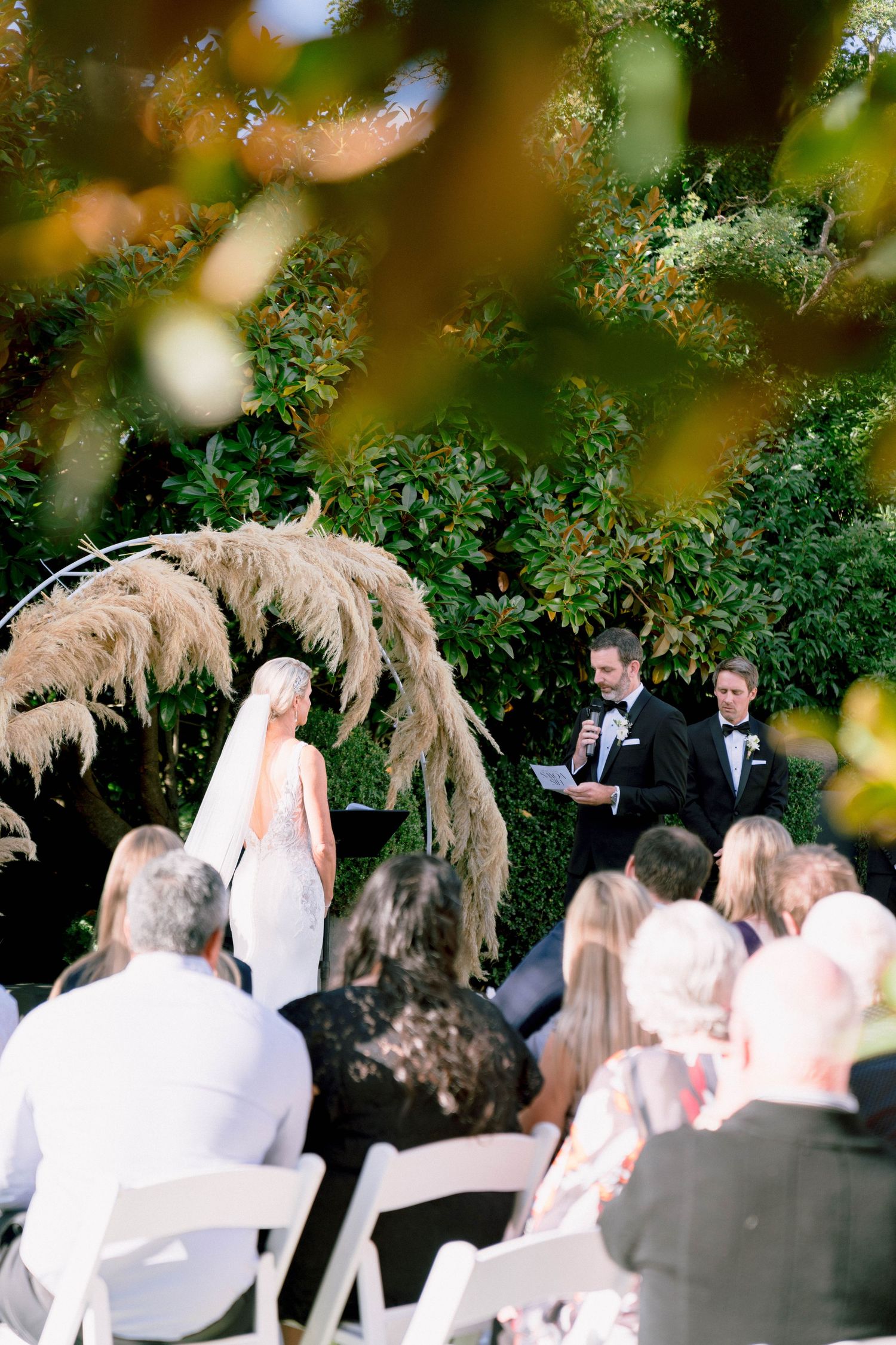 An outdoor wedding ceremony setup with white chairs and floral decorations under trees.