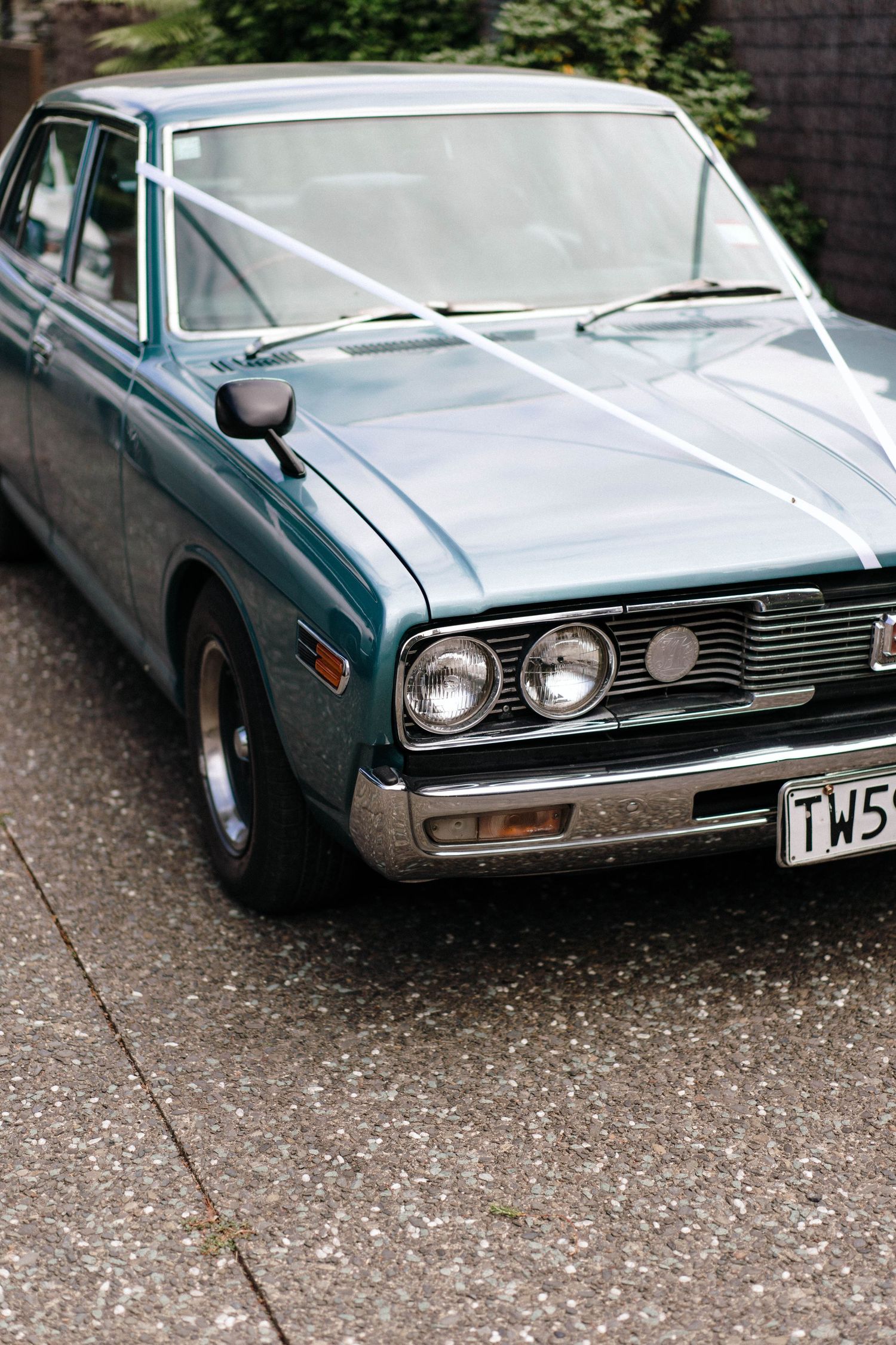 Vintage teal sedan with dual headlights and white ribbon decoration parked on concrete driveway.