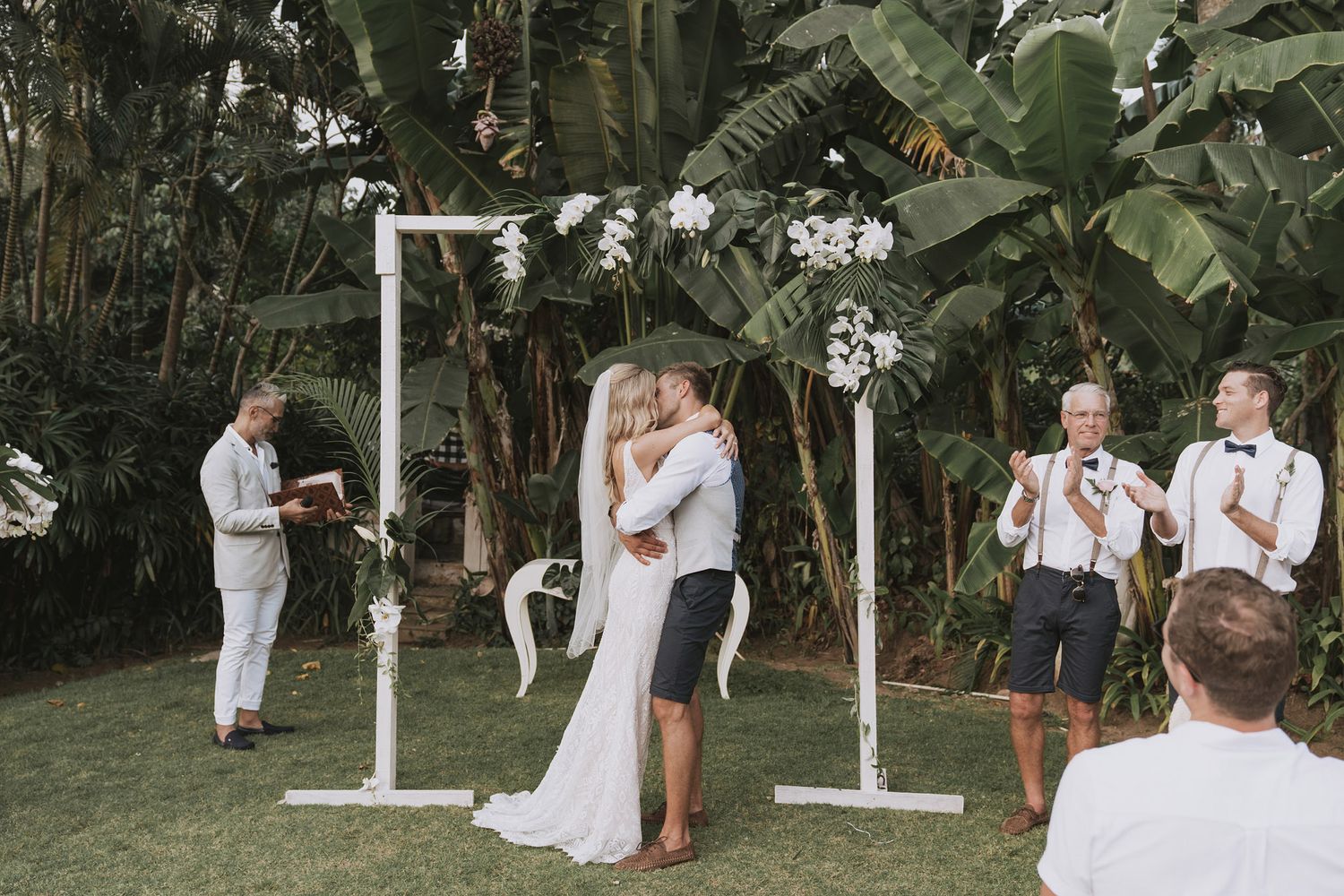 Wedding ceremony takes place under a floral arch on a lush tropical lawn during an intimate outdoor celebration.
