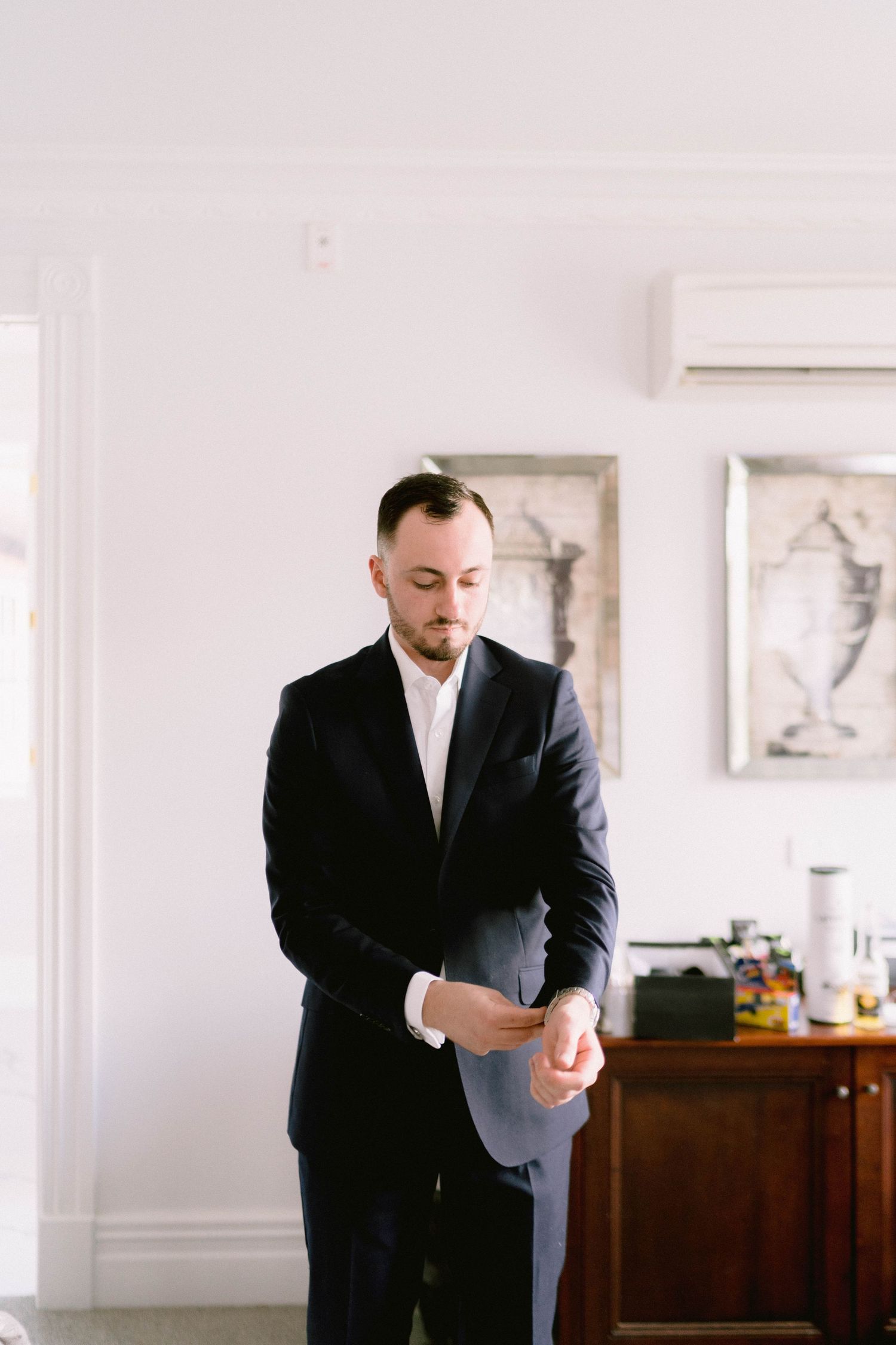 A person in a black suit adjusts their sleeve cuff while standing in a bright room with framed artwork on the wall.