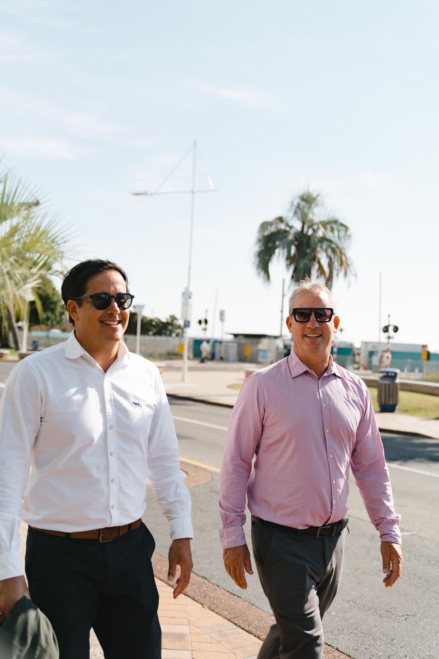 Two colleagues walking along a waterfront path wearing sunglasses on a bright sunny day.