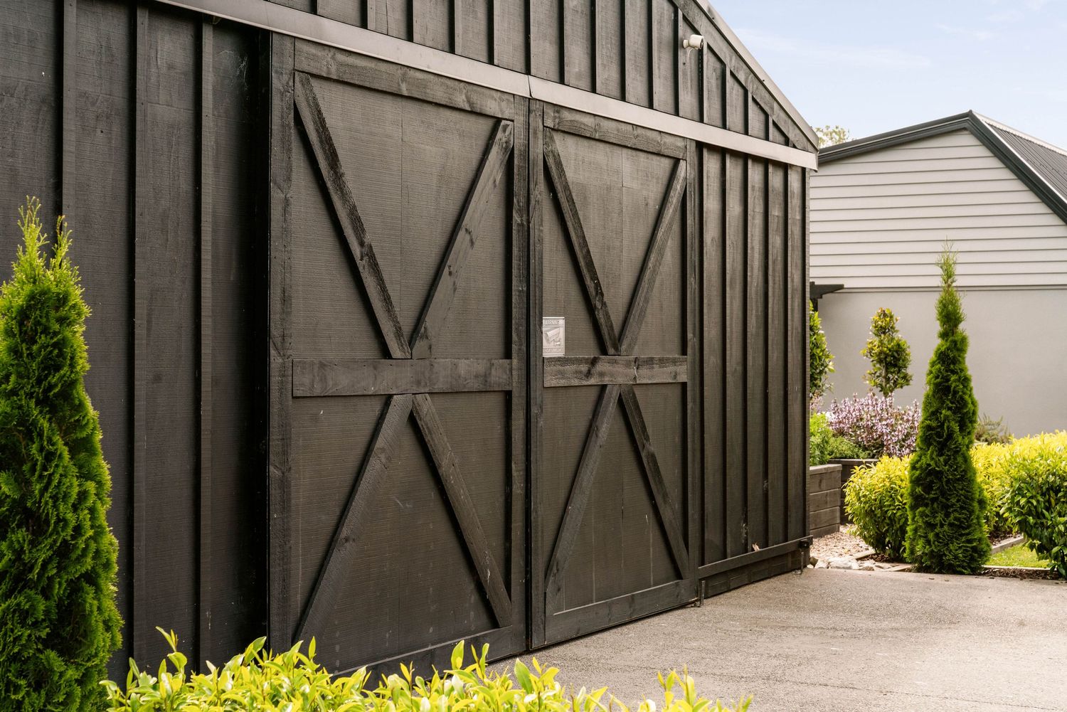 Dark gray modern barn doors with X-patterns are flanked by evergreen shrubs in a well-maintained garden setting.