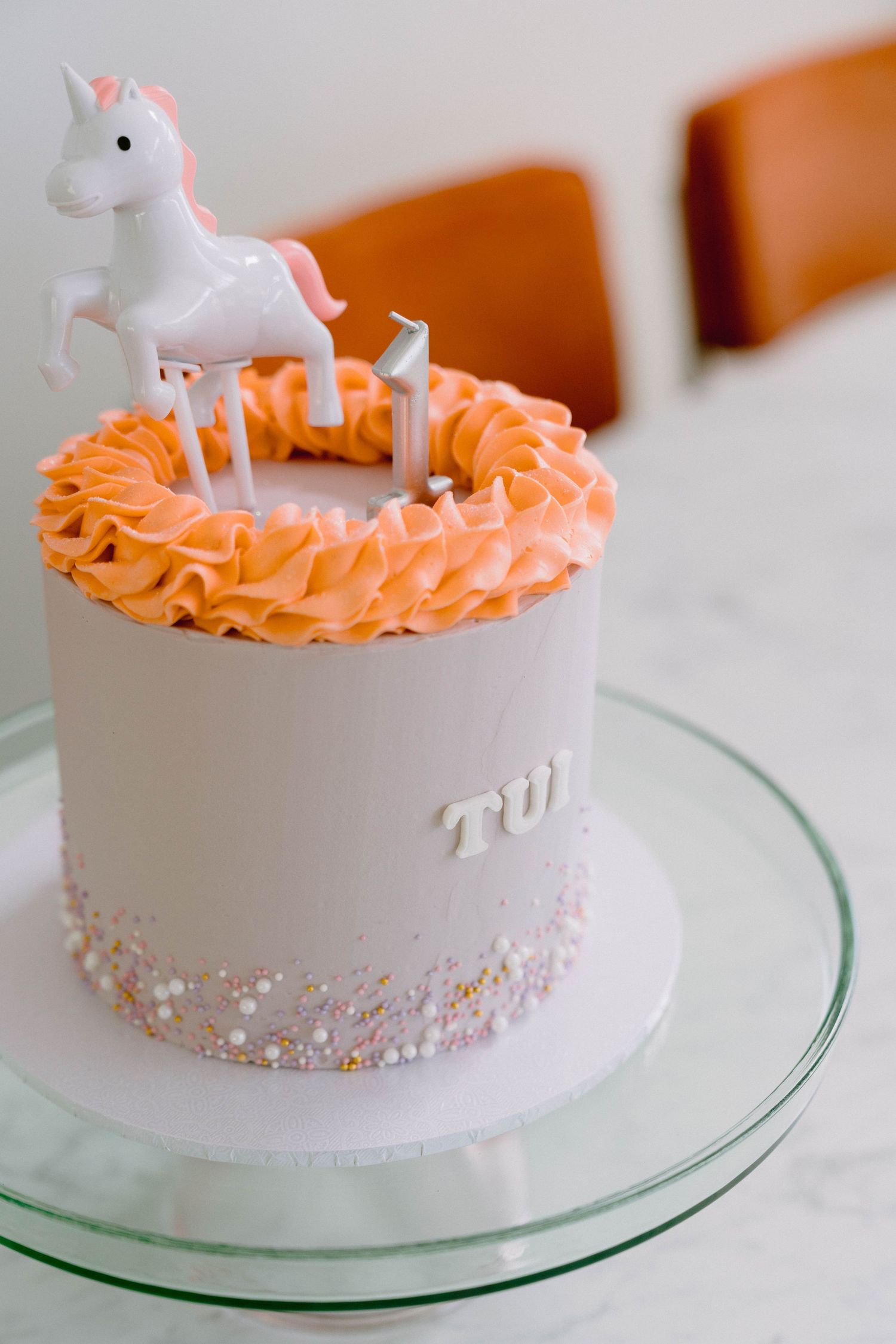 A white birthday cake adorned with pink ruffled frosting and pearl details on a glass cake stand.