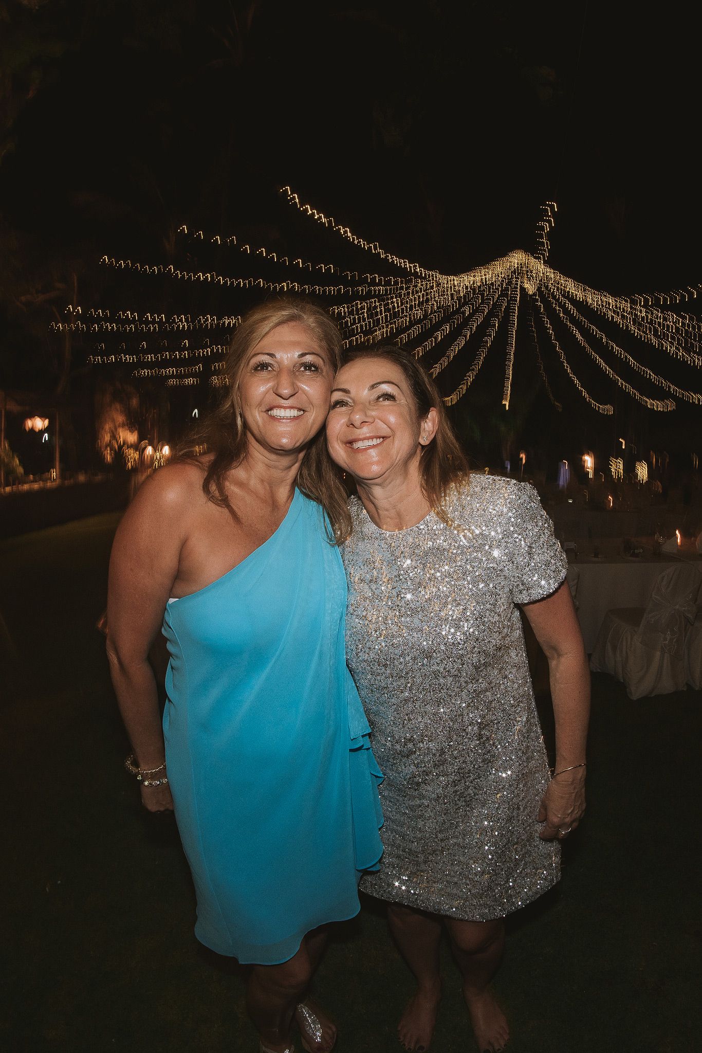 Two guests celebrating at an outdoor evening event under decorative string lighting.