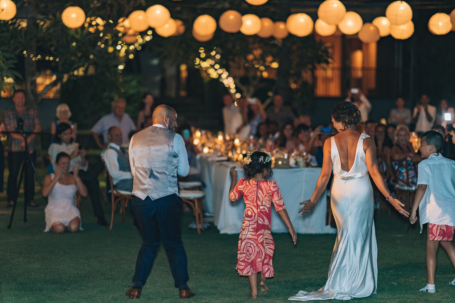 Wedding guests dance on a lawn decorated with glowing lanterns during an outdoor evening reception.