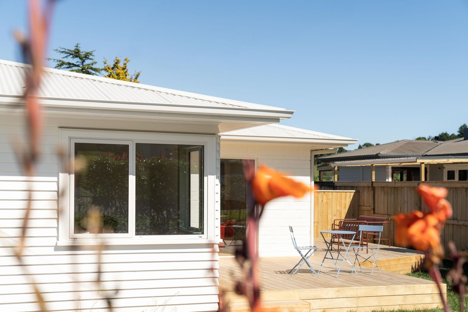 White single-story house with large window and patio furniture viewed through orange flowers against blue sky.