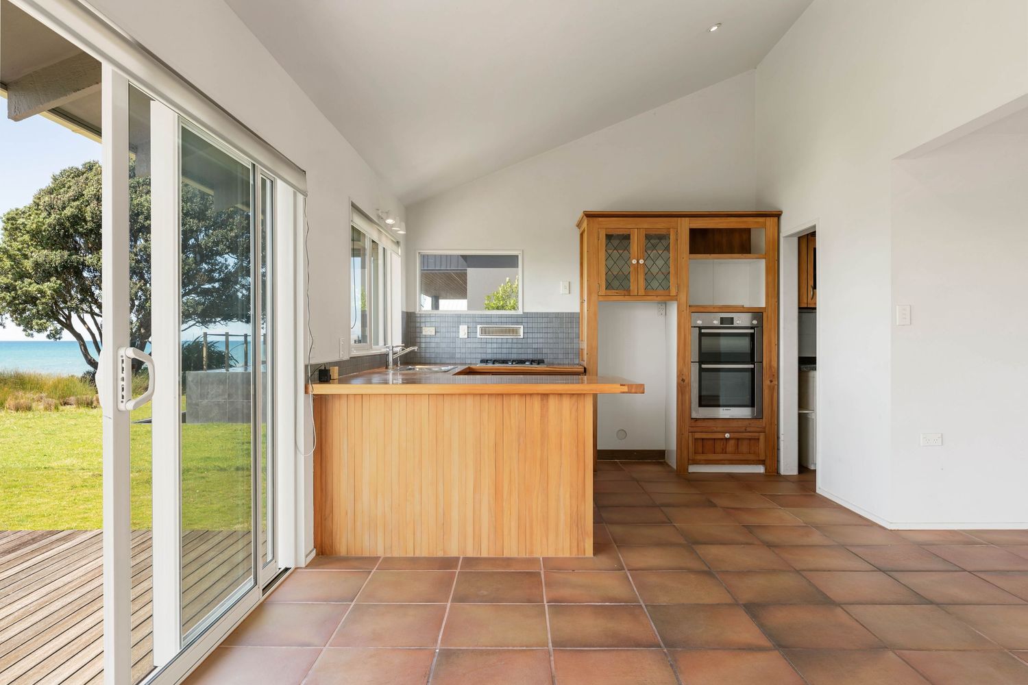 Modern kitchen with wooden cabinetry, glass sliding doors and terracotta tile flooring overlooking an outdoor view.