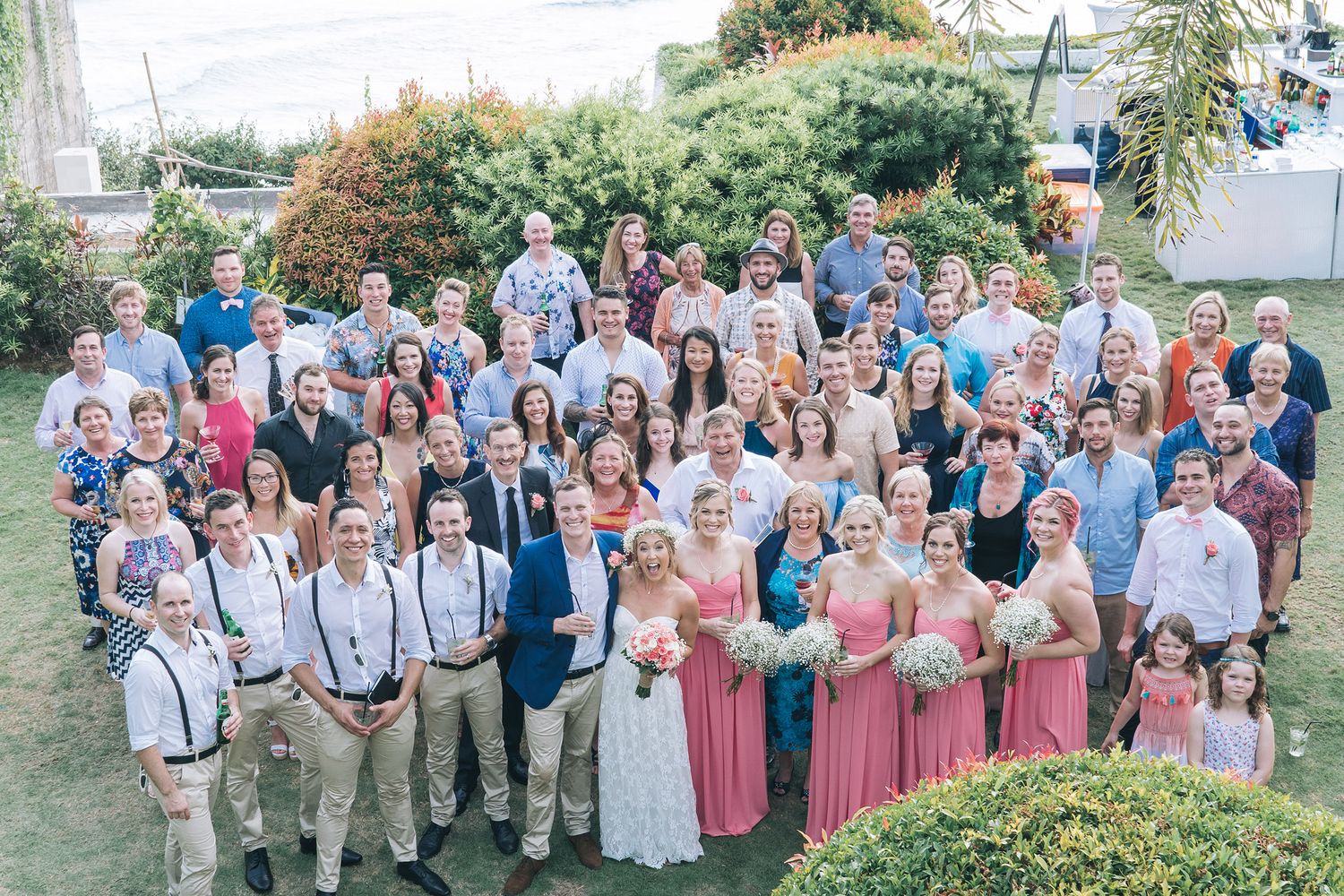 Large group wedding photo with attendees in coordinated pink and navy attire on a grassy hillside.