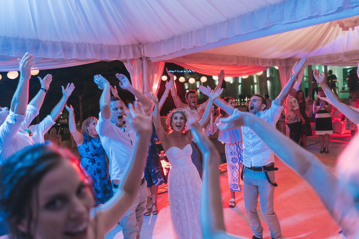 Wedding guests dancing and celebrating under tent with colorful lighting.