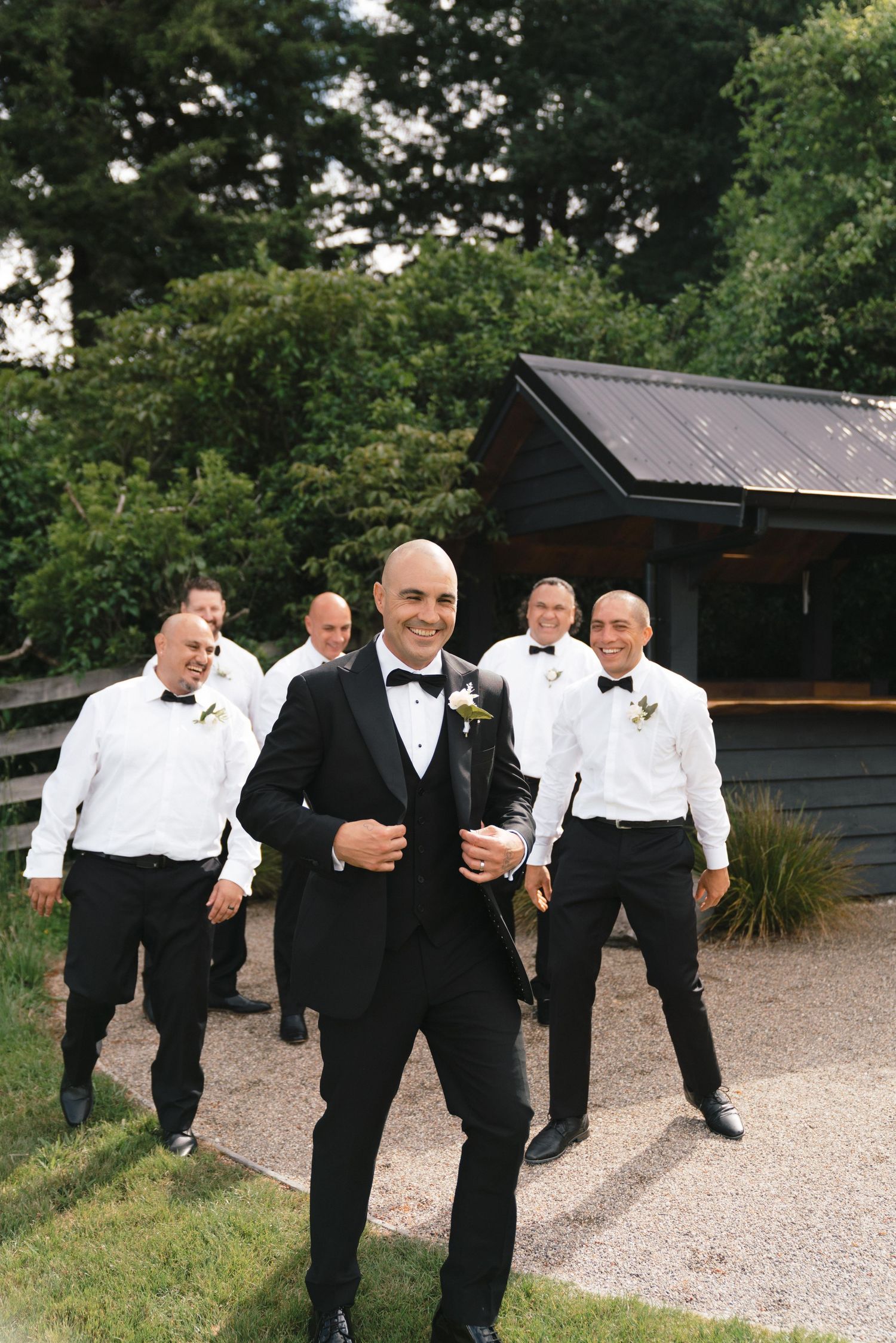 Groomsmen in black tuxedos and bow ties walking and posing playfully on a gravel path outdoors.