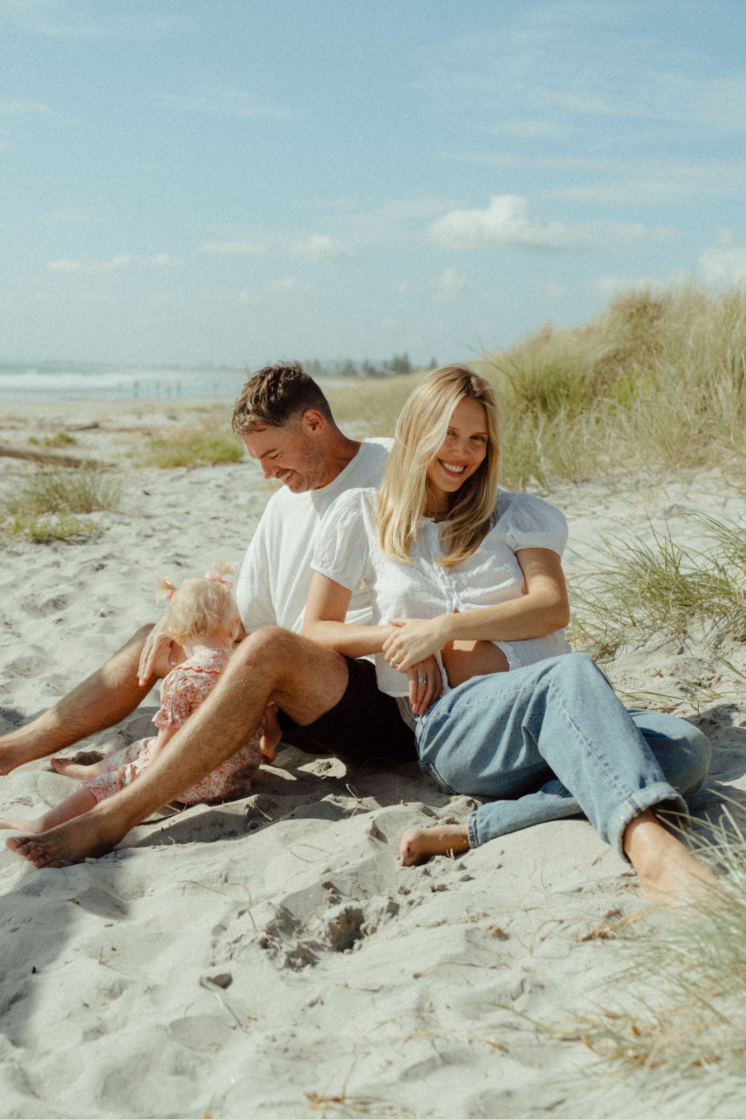 A couple relaxes together on a sandy beach with dunes and blue sky on a sunny summer day.