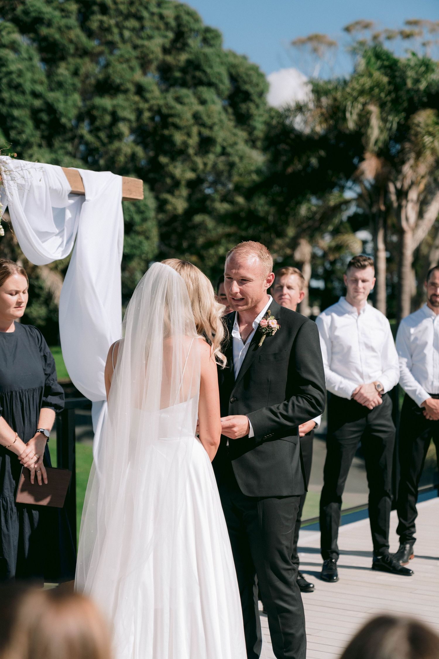 Wedding couple shares a romantic moment with wedding party on an outdoor walkway lined with palm trees.
