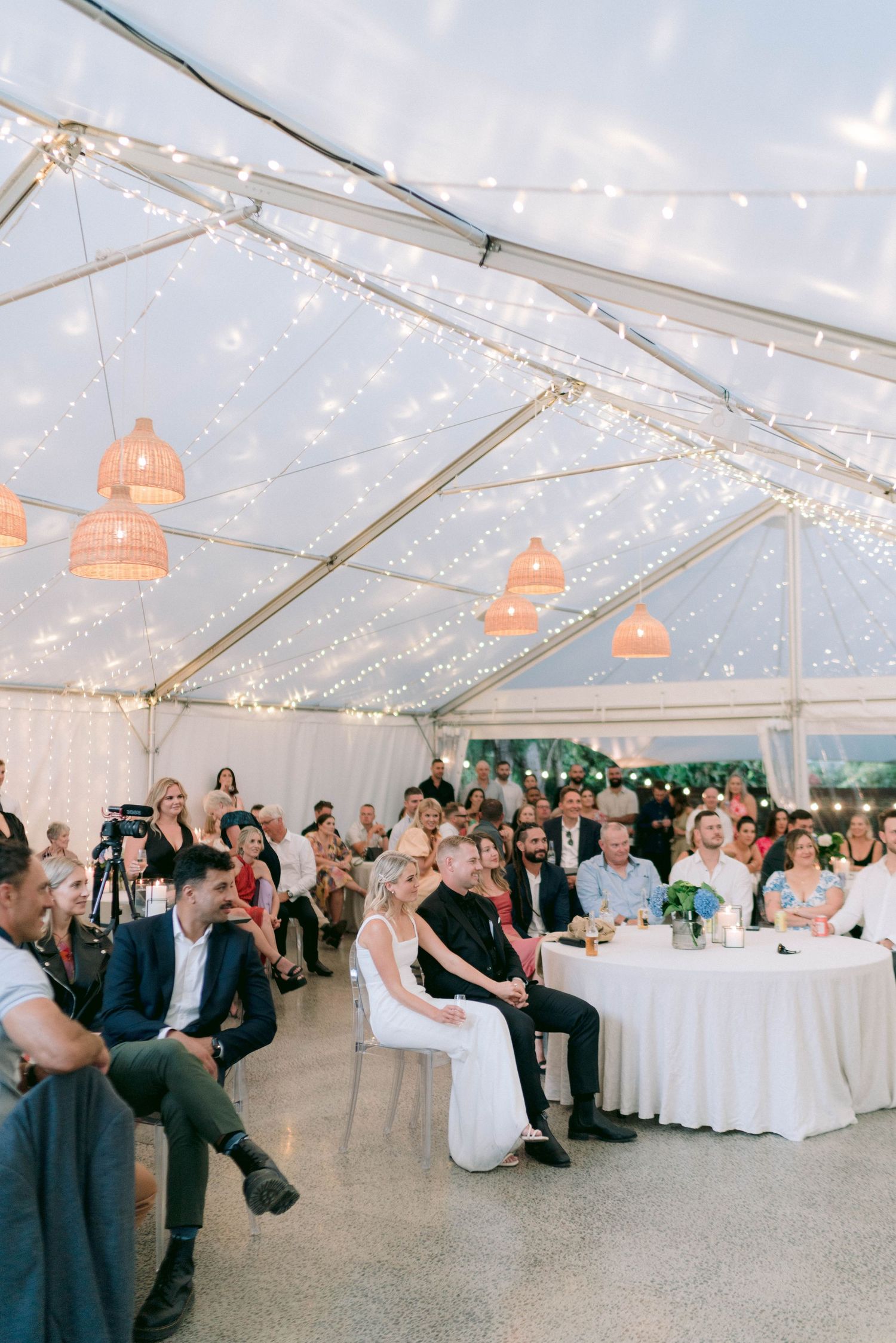Reception celebration in clear tent decorated with string lights and coral pendant lamps above dining tables.