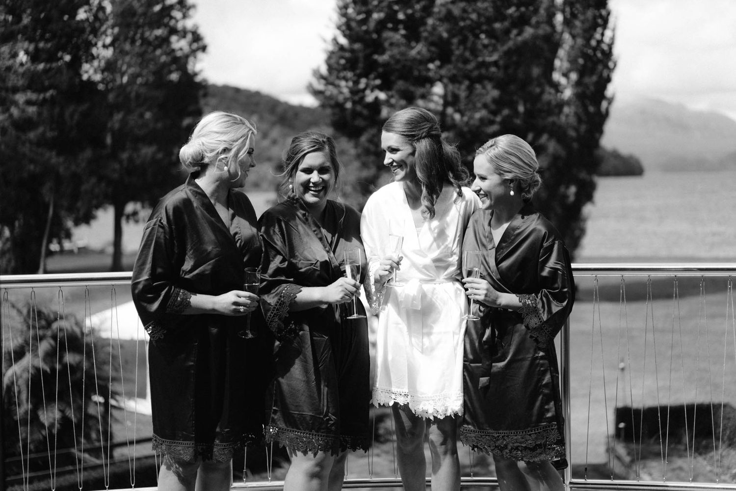 Vintage black and white photo of four fashionably dressed women chatting outdoors with a lake and trees in background.