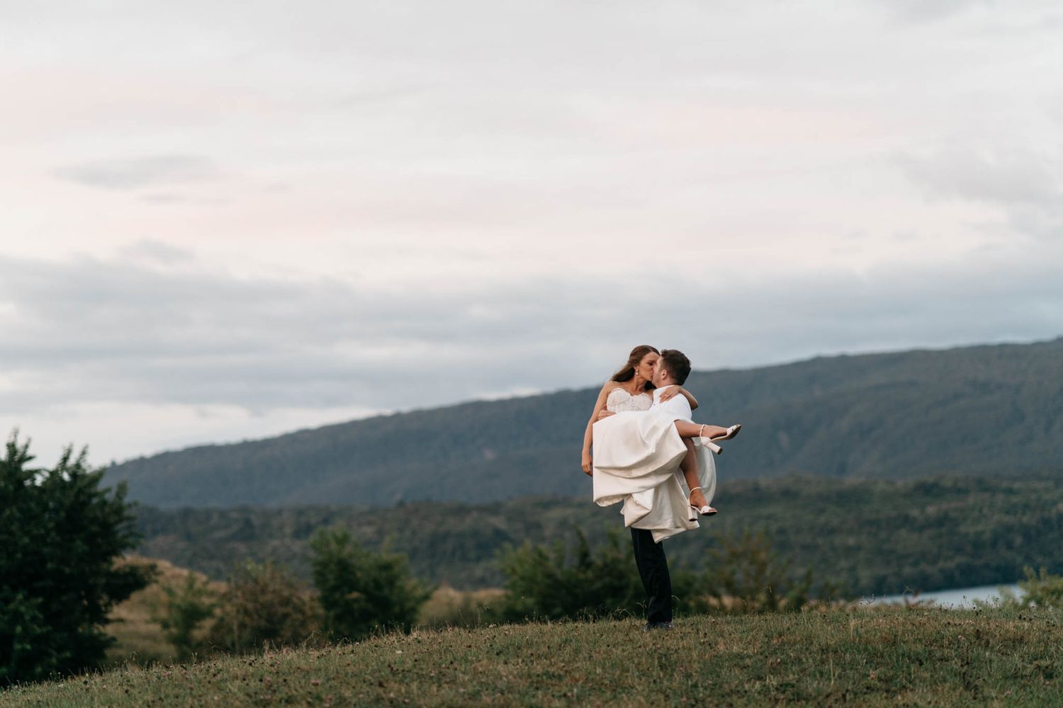 A couple stands intimately together on a grassy hilltop overlooking a scenic mountain landscape during sunset.