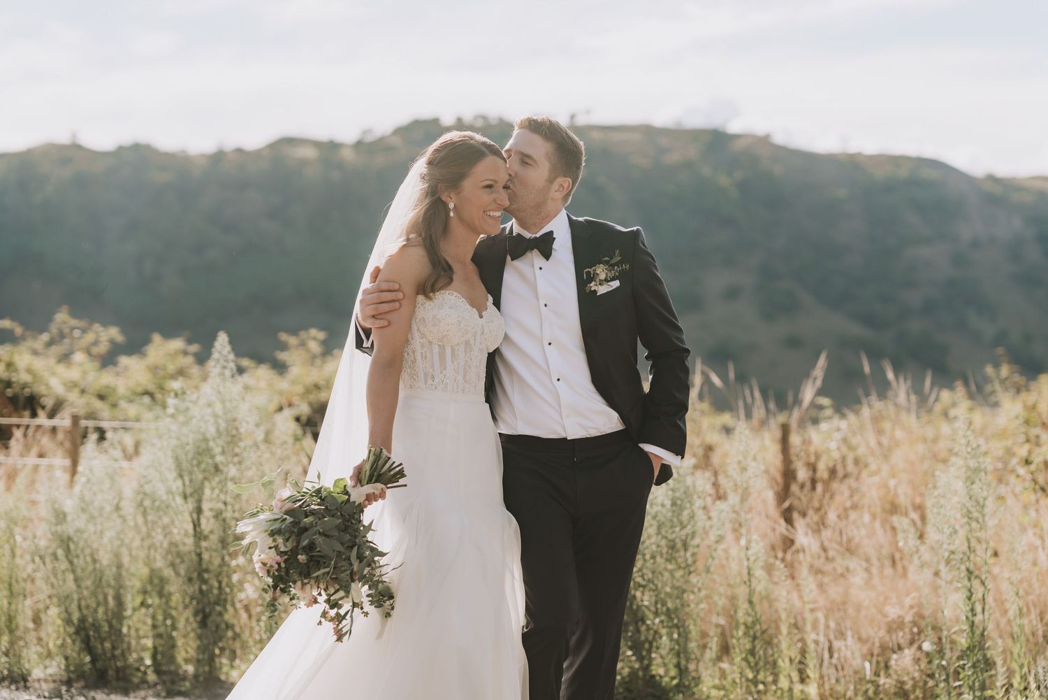 Newlywed couple stands close together in a field of tall grass during golden hour at their outdoor wedding.