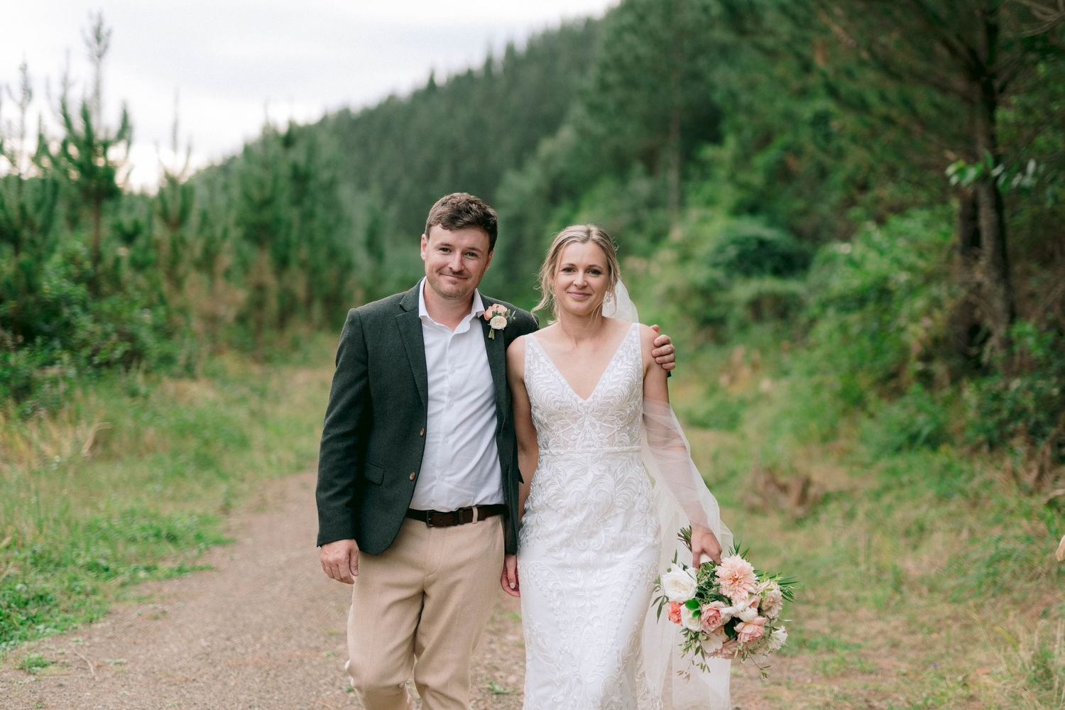 A couple walks together on a forest path while wearing wedding attire and holding a floral bouquet.