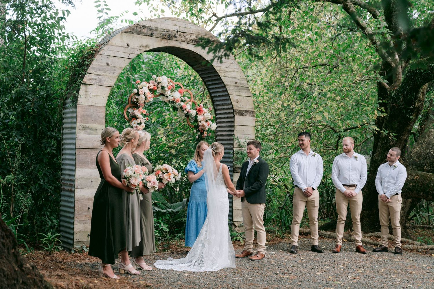 Wedding party poses for photos under a decorative stone arch in a lush garden setting.