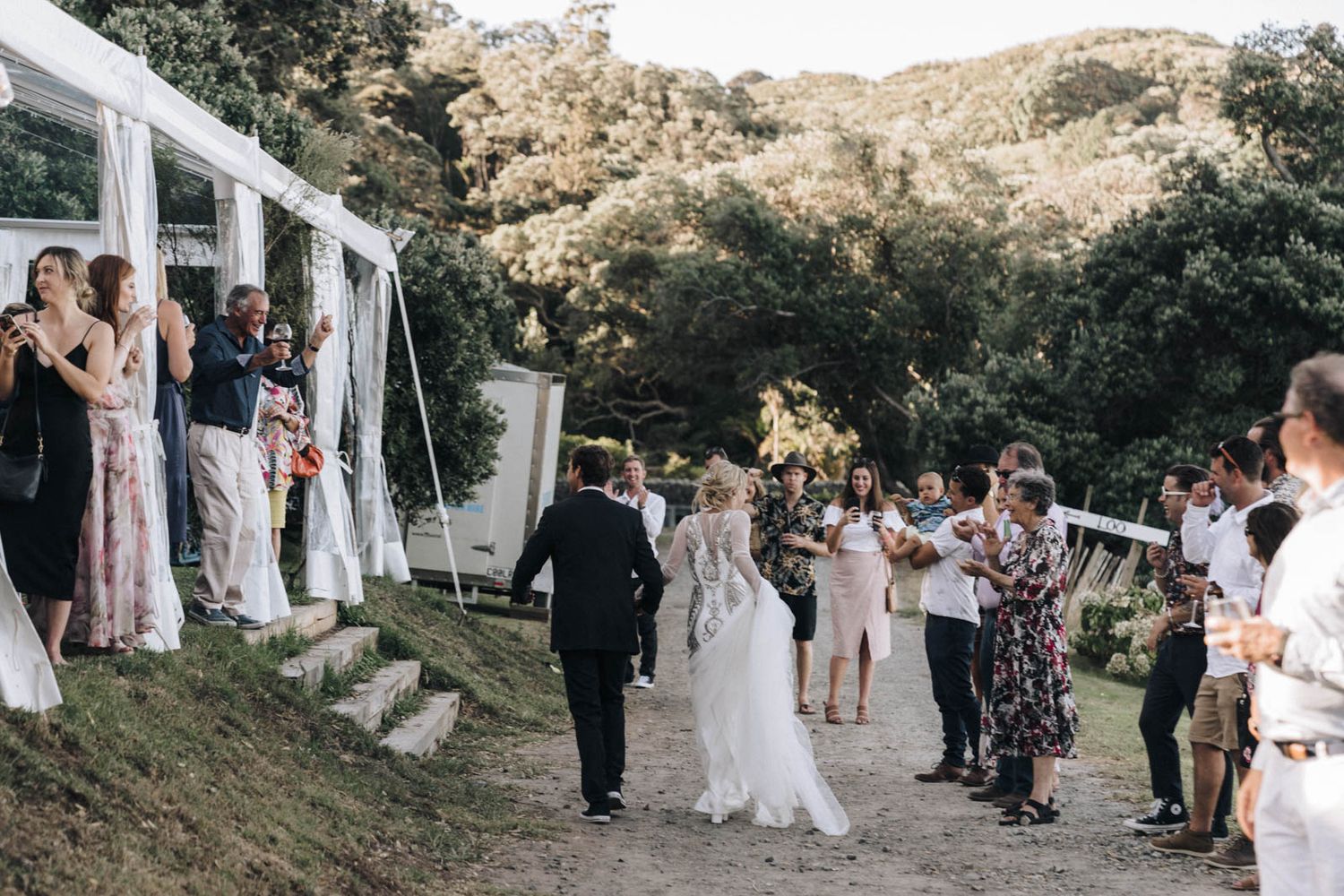Wedding celebration on a hillside with guests lining pathway under white tent awnings during an outdoor ceremony.
