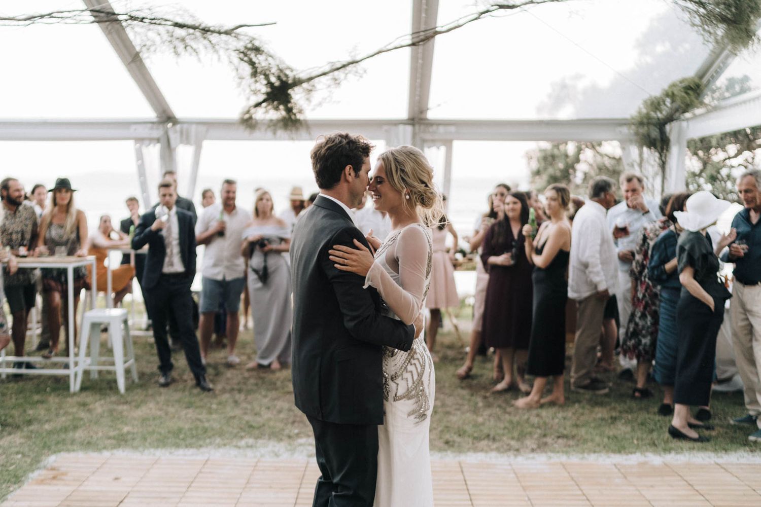 Couple shares their first dance under a white tent while wedding guests watch from the perimeter at an outdoor reception.