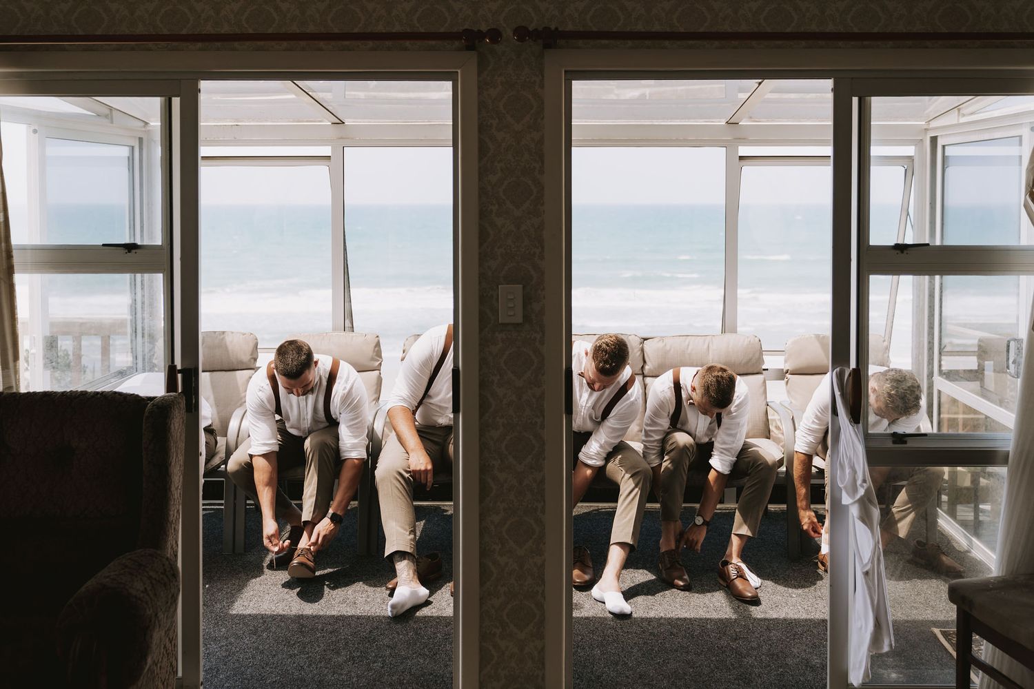 Wedding party in suspenders and formal wear sits by oceanfront windows during preparation moments.