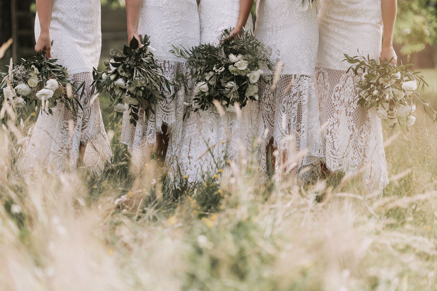 Wedding party members in white lace dresses hold elegant greenery bouquets while standing in tall grass.