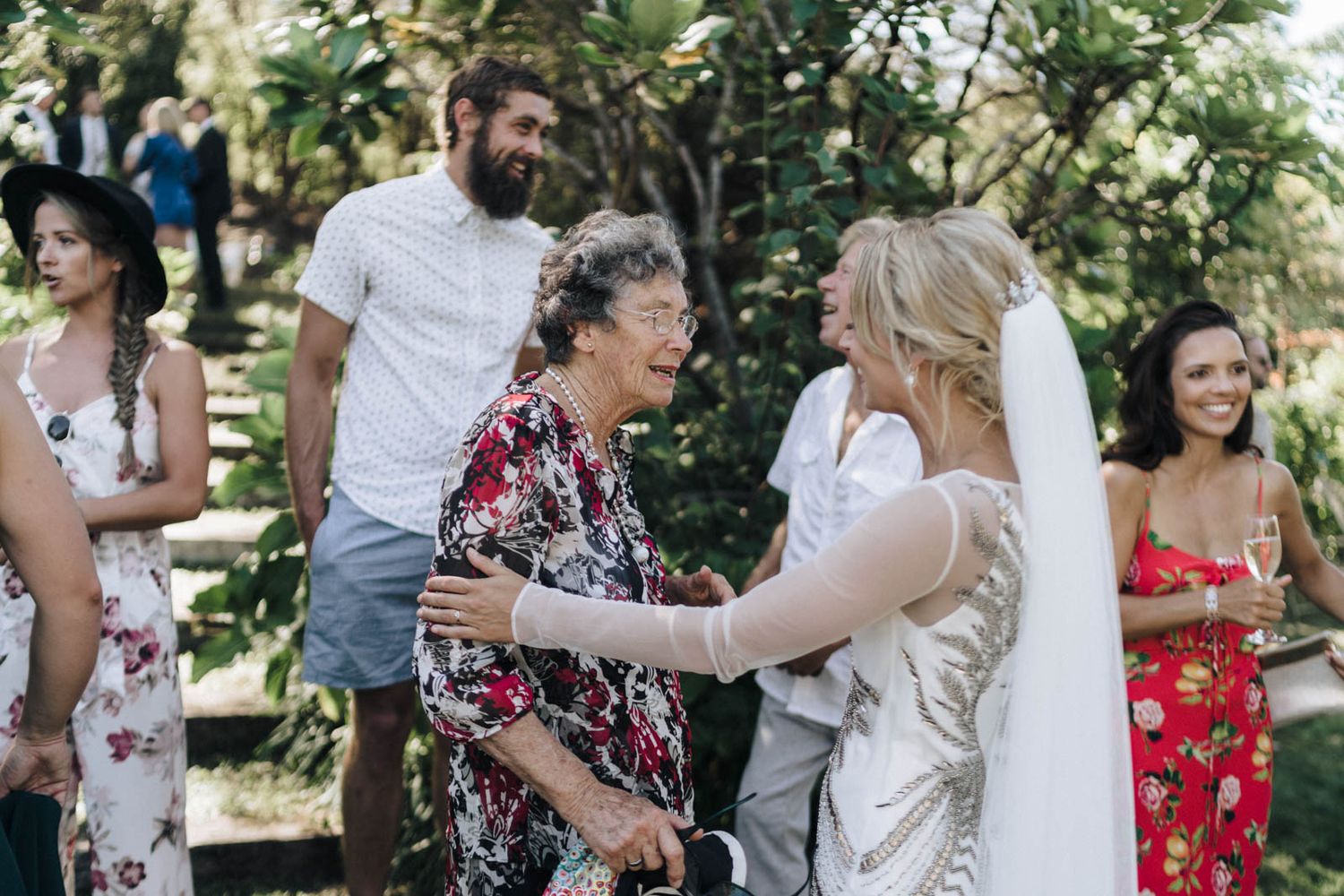 Joyful wedding celebration in a garden setting with guests in casual summer attire sharing embraces and laughter.