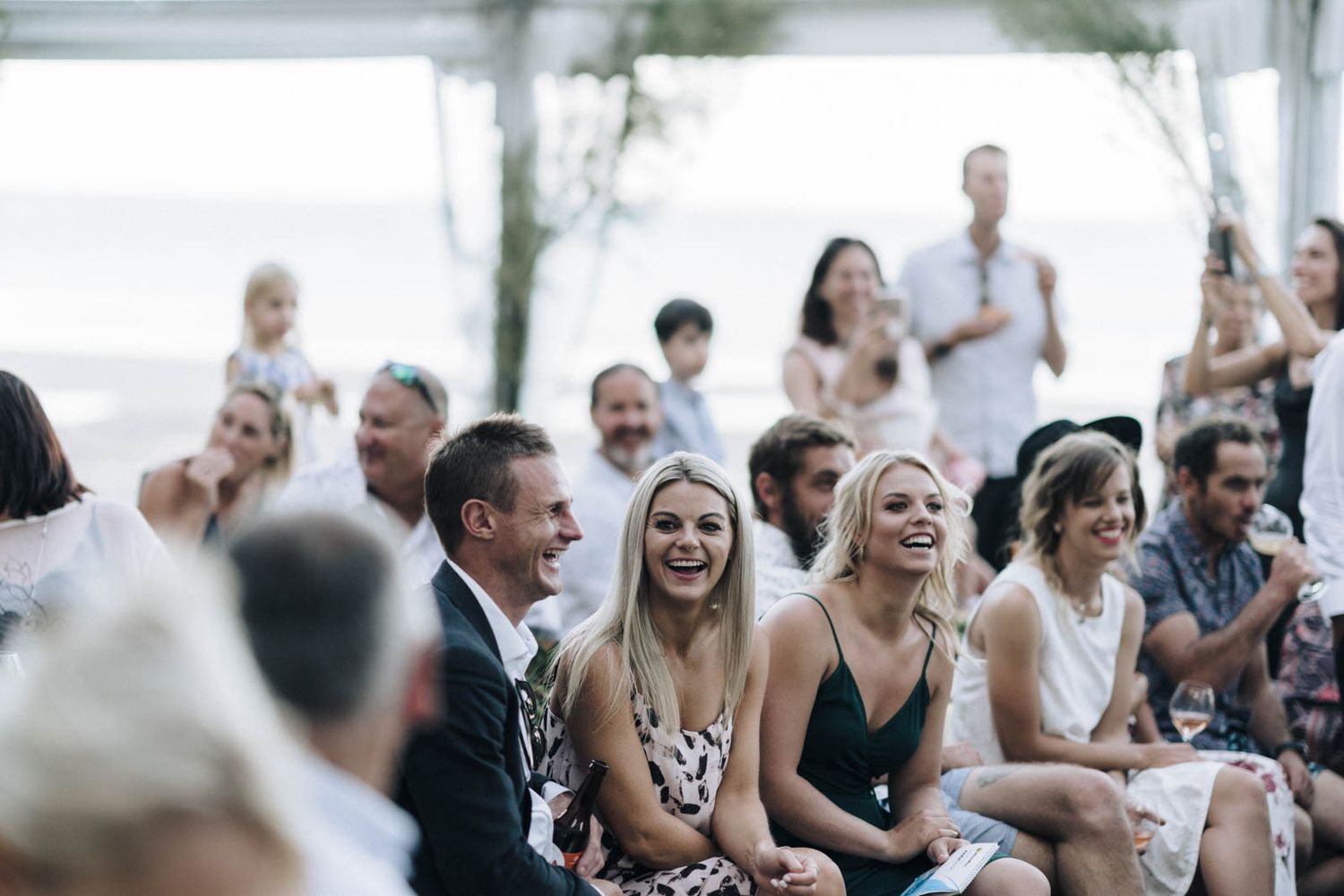 Wedding guests smile and laugh while seated outdoors under a white tent during a summer ceremony.