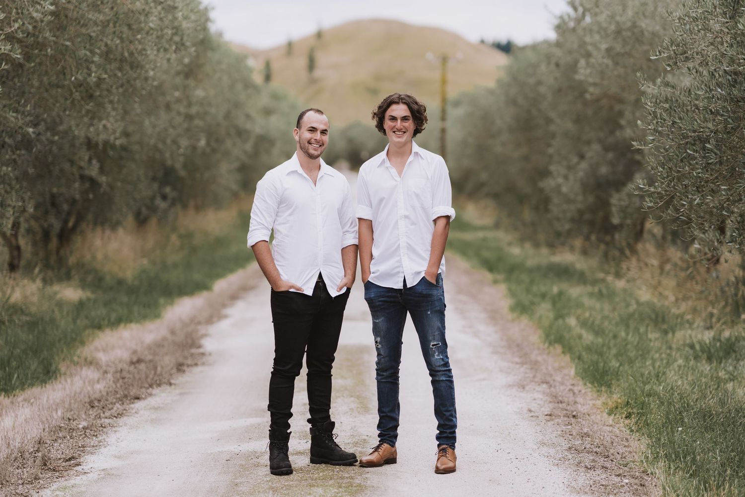 Two people in matching white shirts and dark pants walk together on a rustic dirt path surrounded by olive trees.