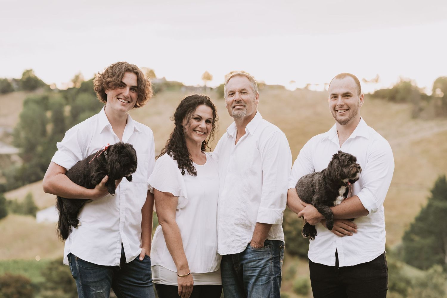 Group in white shirts pose together on a hill at sunset while holding black puppies.