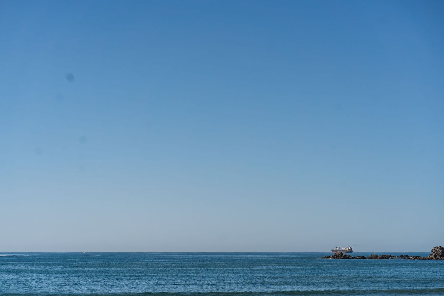 Calm blue ocean meets clear sky at the horizon with rocky outcrops visible in the distance.