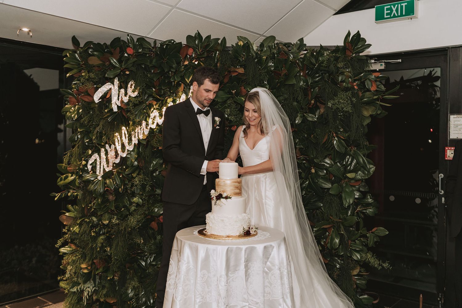 A bride and groom pose with their wedding cake in an elegant venue decorated with greenery and floral arrangements.