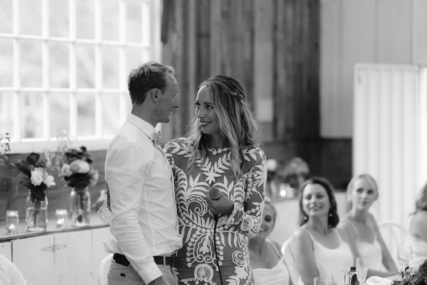 A couple shares a joyful moment in black and white at a wedding reception with guests looking on and floral decor.