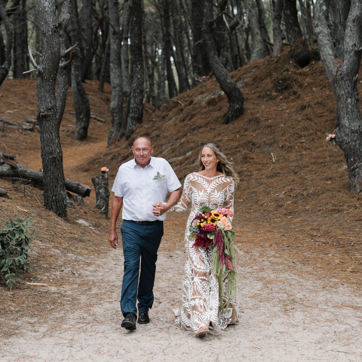 A couple walks together on a dirt path through a forest while one holds a vibrant autumn-colored bouquet.