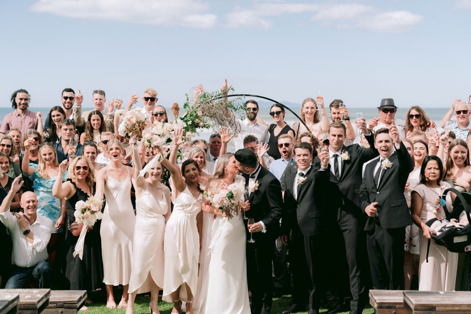 Large group of wedding guests gathered outdoors for a formal group photo by the ocean.