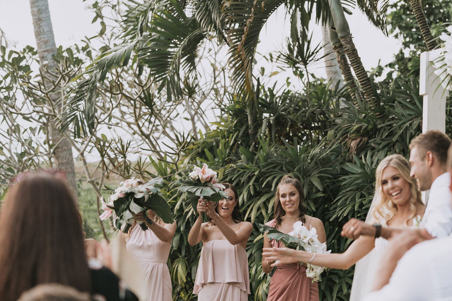 Wedding guests dance outdoors at a tropical venue with palm trees during a lively reception celebration.