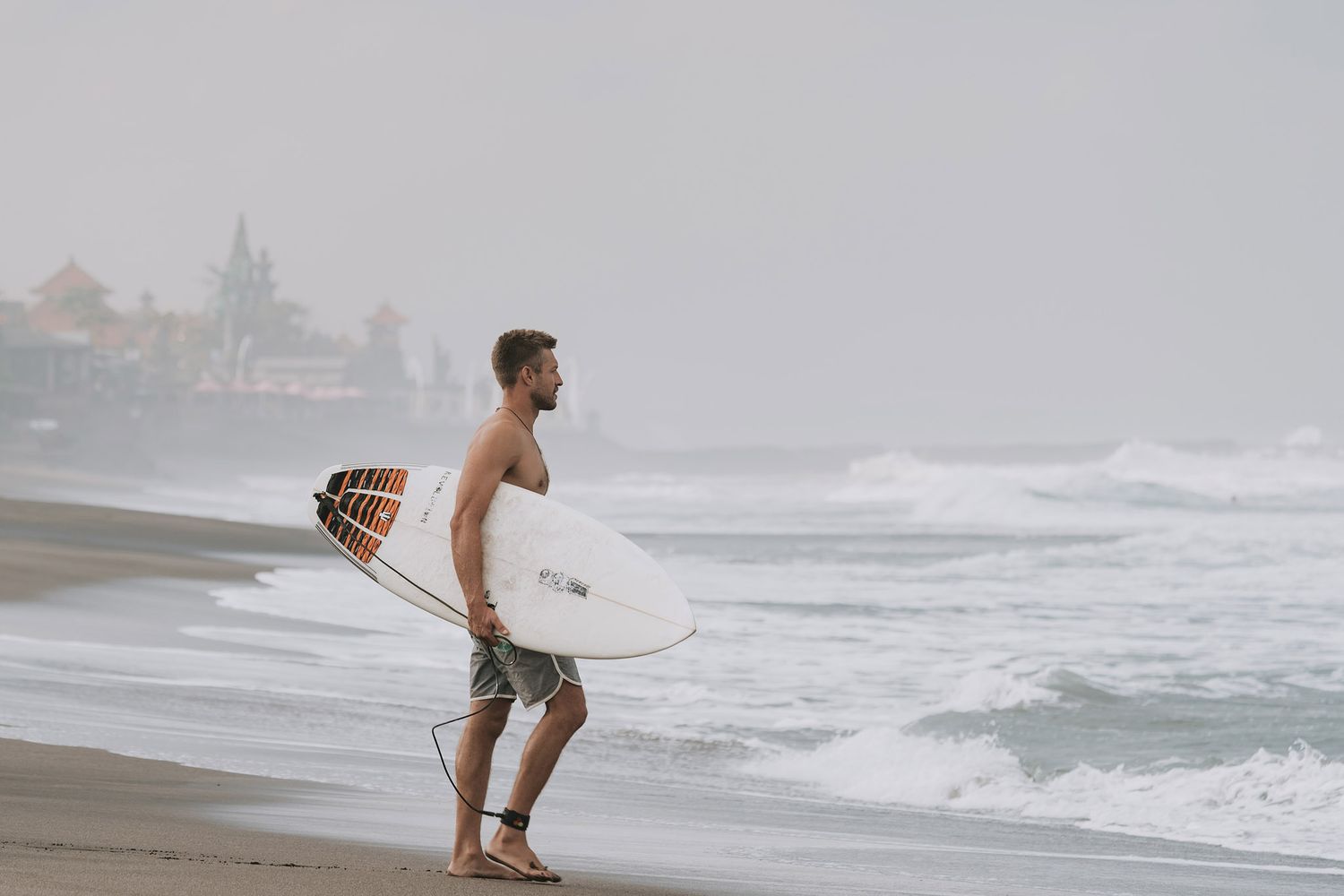 Surfer holding white surfboard stands on misty beach during stormy conditions with waves crashing in background.
