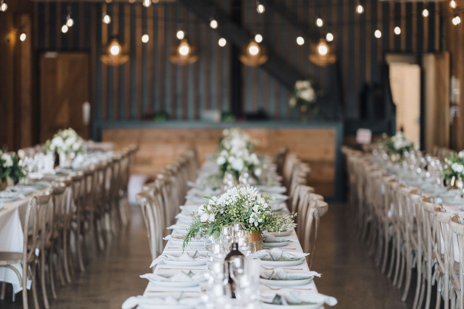 Elegant rustic wedding reception setup with long tables, white linens and hanging lights in a wooden barn venue.