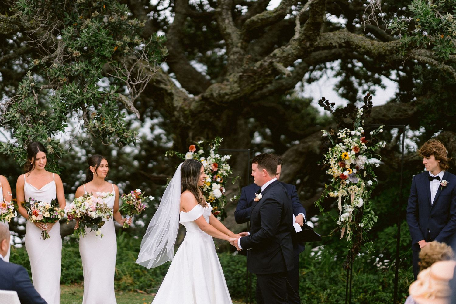 Wedding ceremony taking place under a large tree with floral arrangements and bridesmaids.