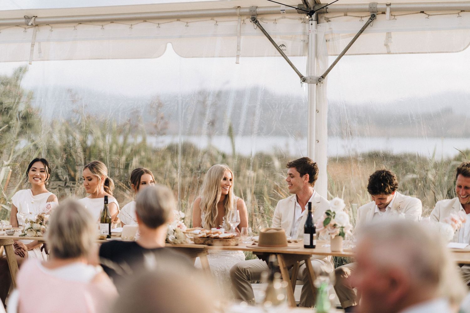 Wedding guests gather under a white tent for an outdoor reception with water views.