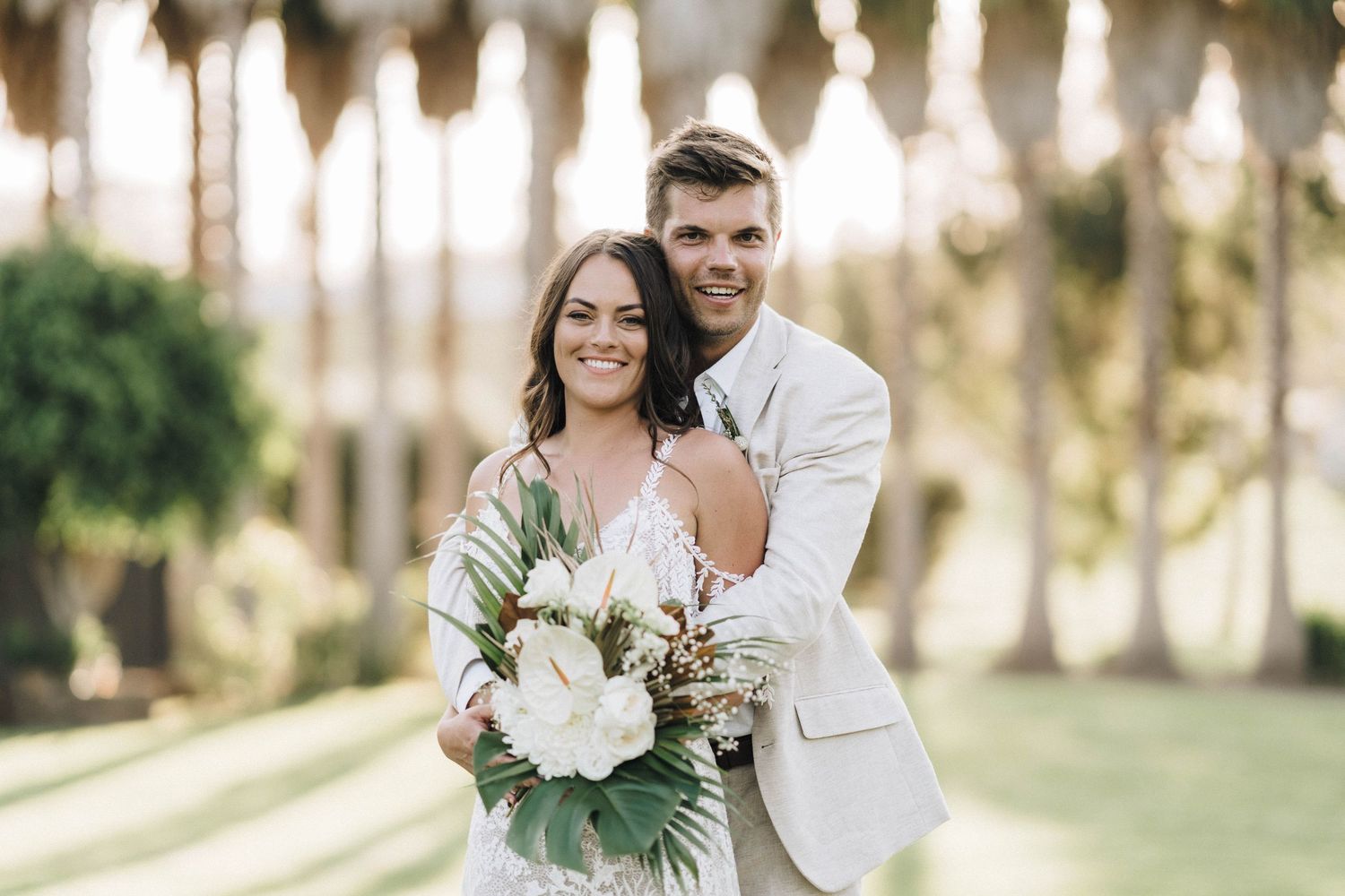 Elegant wedding portrait with couple holding tropical bouquet under palm trees.